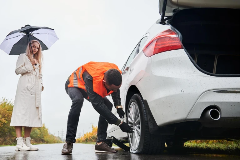 Technician Inspecting a Flat Tyre on the Roadside with a Car Owner Nearby